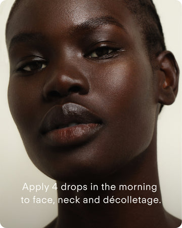 Close-up of a woman with dark skin looking at the camera. Text reads: Apply 4 drops of C+ Rejuvenating C Chebula Facial Serum Mini in the morning to face, neck, and décolletage.