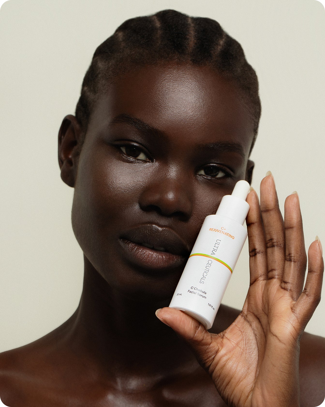 A person with dark skin and short, braided hair holds a C+ Rejuvenating C Chebula Facial Serum Mini bottle with a dropper near their face against a light background.