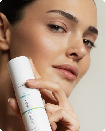 A woman with clear, glowing skin holds a white bottle of B² Hydrating HA 360 Facial Serum Mini near her face, highlighting the product. She wears natural makeup and has a soft, confident look.