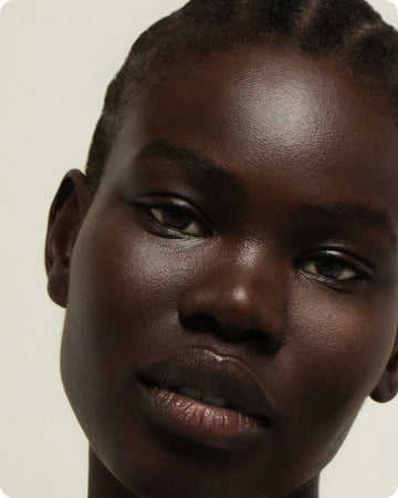 A close-up portrait of a person with dark skin and braided hair, posing against a light background that highlights their smooth, radiant complexion enhanced by Ultra C Firming Concentrate.