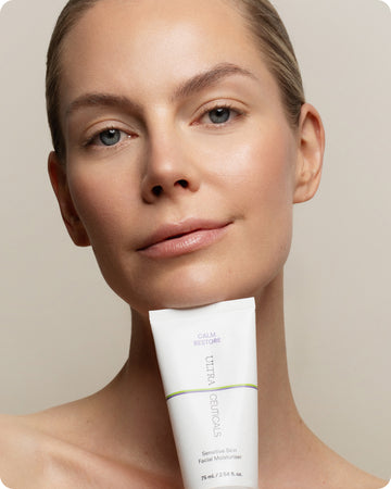 A woman with clear skin smiles softly, holding a white tube of Ultra Calming Moisturiser Cream under her chin against a neutral background, highlighting its gentle formula for sensitive skin.