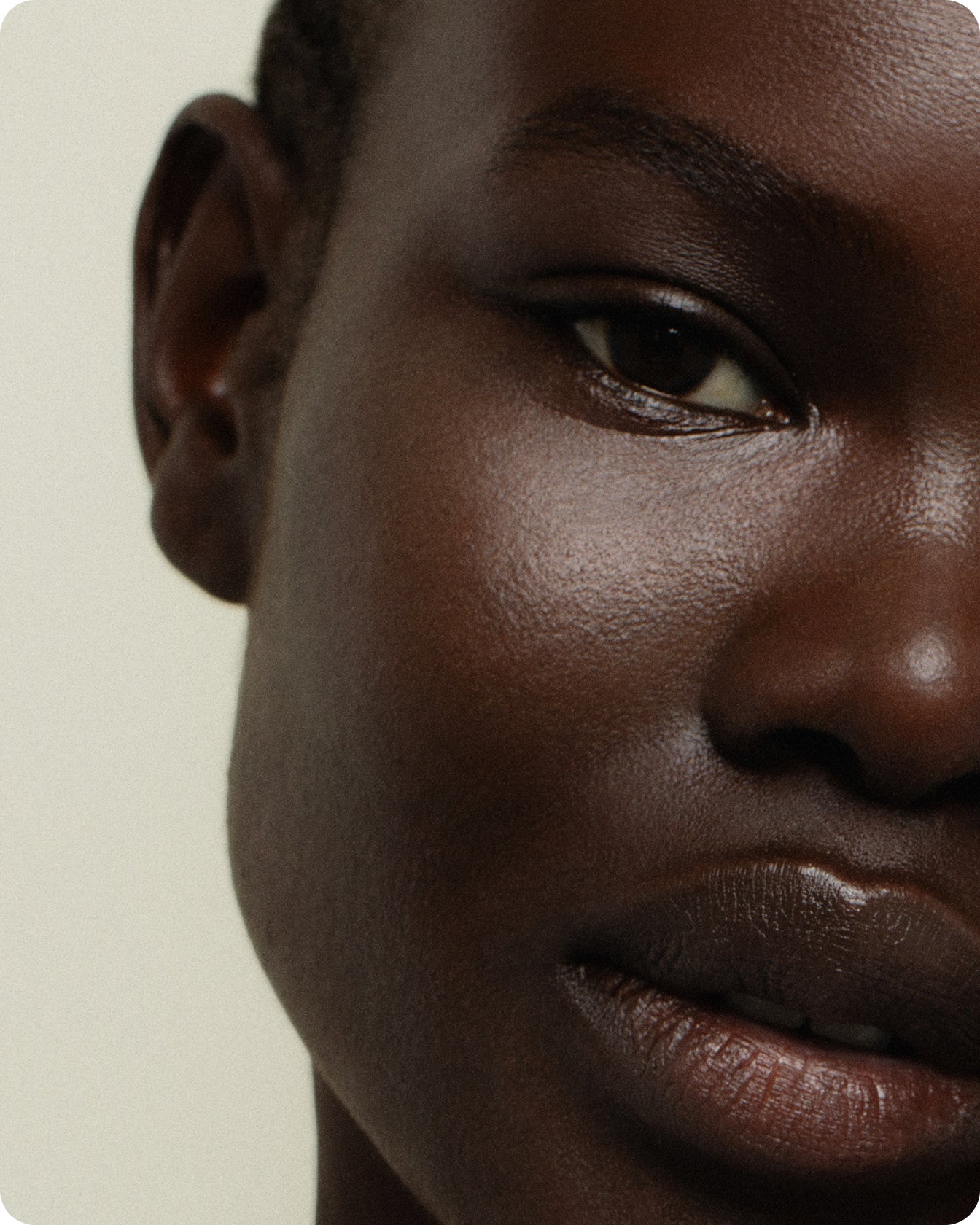 Close-up of a person's face with dark skin, showcasing detailed texture enhanced by C+ Rejuvenating Pro-Collagen Night Cream. Soft lighting highlights the eye, eyebrow, nose, lips, and ear against a neutral background.