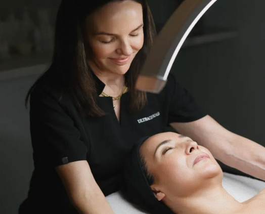 A beauty therapist in a black uniform with 'ULTRACUSHION' branding smiles while treating a client lying down under a curved light in a spa setting.