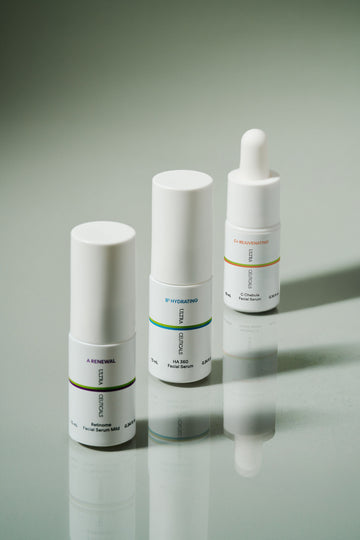 Three white skincare serum bottles with minimal labels stand upright on a reflective surface. The ABC Mini Essentials kit features a bottle with a dropper cap and colored text accents, perfect for sampling Vitamin C and retinol serums.