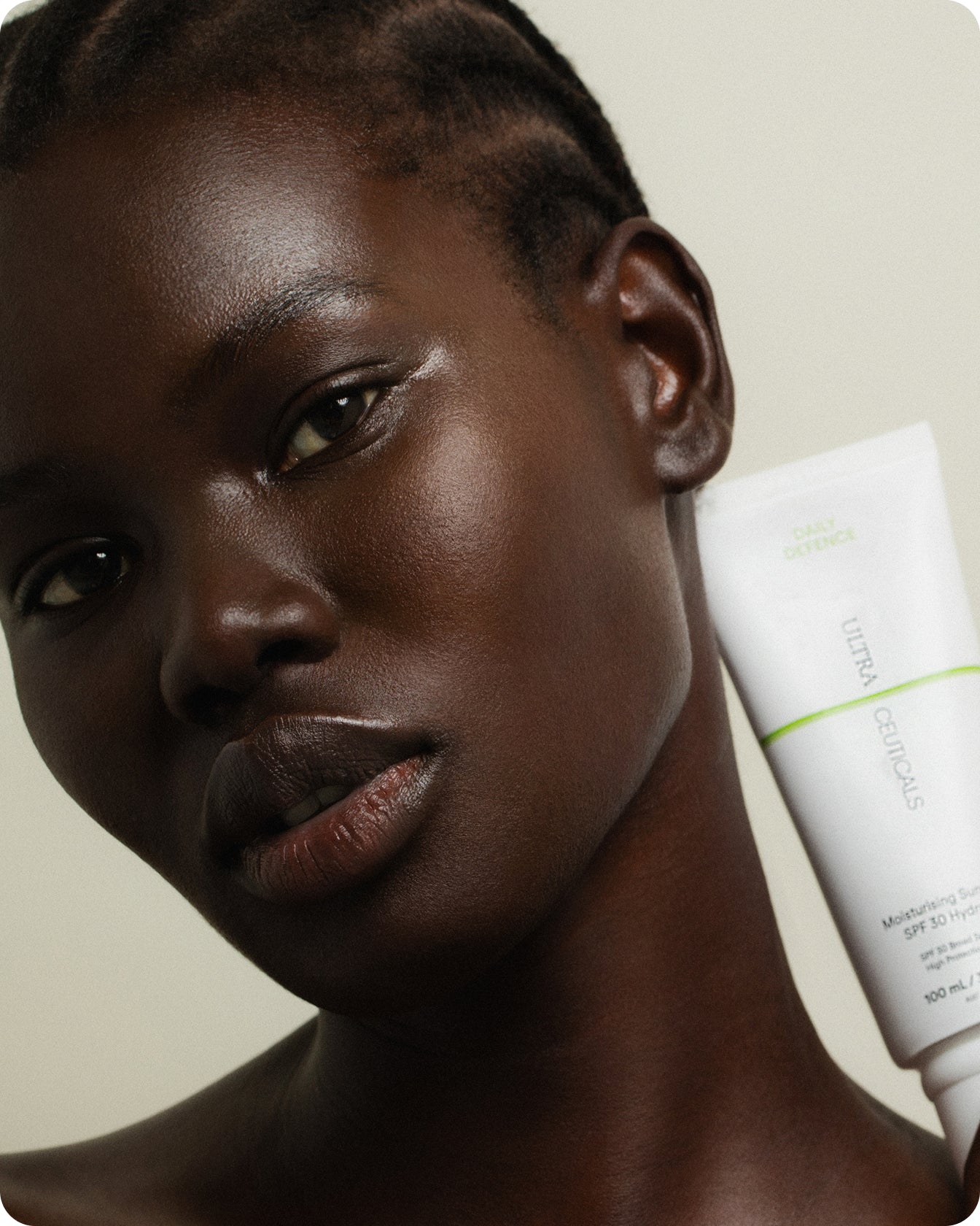 A person with dark skin and short braids holds a white tube of Ultra UV Protective Daily SPF30 Sheer Tint near their face, looking at the camera against a plain, light background.