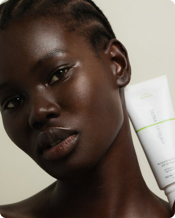 A person with dark skin and short braids holds a white tube of Ultra UV Protective Daily SPF30 Sheer Tint near their face, looking at the camera against a plain, light background.