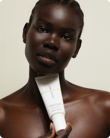 A woman with dark skin and short hair holds a white tube labeled Ultra UV Protective Daily SPF50 Hydrating under her chin, looking at the camera. This SPF moisturiser provides hydration and sun protection against a neutral background.