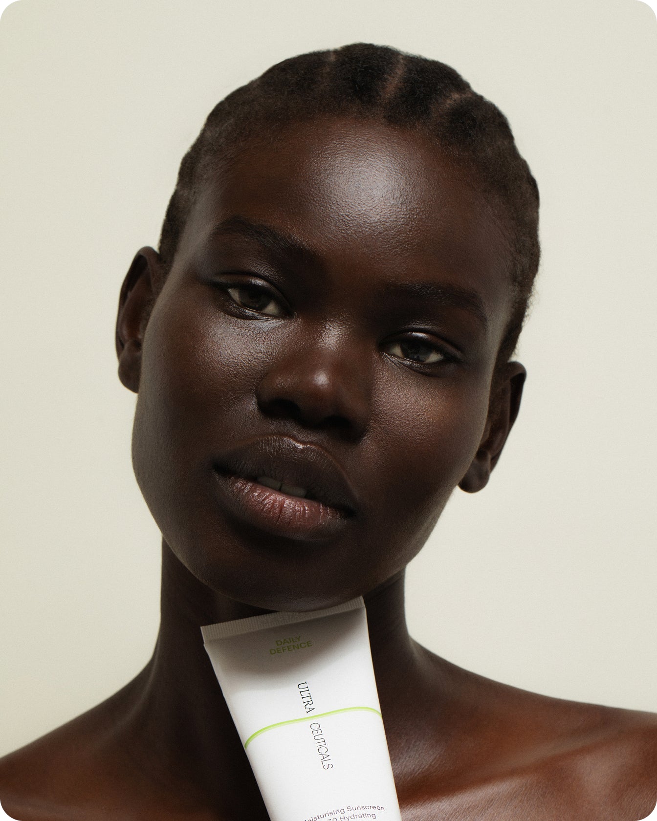 A woman with dark skin and braided hair holds a tube of Ultra UV Protective Daily SPF50 Mattifying sunscreen under her chin, looking directly at the camera against a plain background.