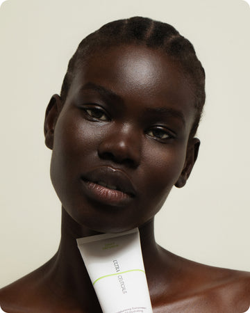 A woman with dark skin and braided hair holds a tube of Ultra UV Protective Daily SPF50 Mattifying sunscreen under her chin, looking directly at the camera against a plain background.